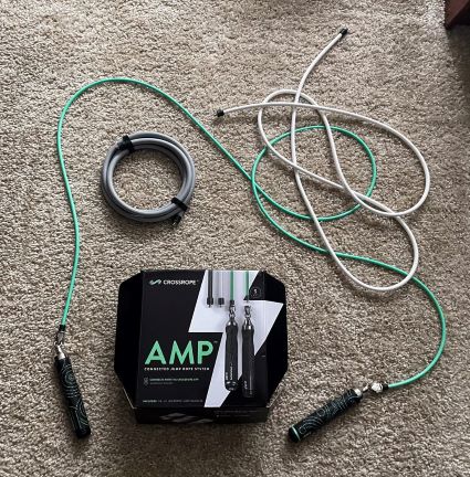 Crossrope AMP 跳绳套装盒周围环绕着三根绿色、灰色和白色加重跳绳，AMP 手柄连接到绿色一根