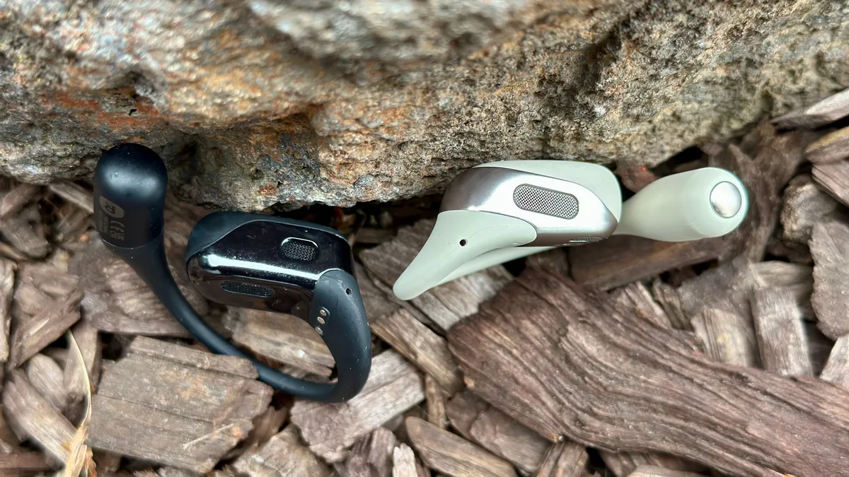 黑色 Shokz OpenFit 和白色 Shokz OpenFit 2 耳塞的俯视图，突出了旧款耳塞上的扬声器和凸起的充电点。