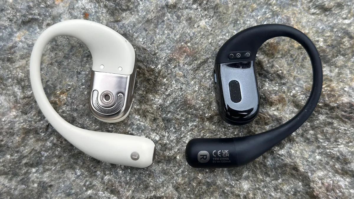 白色 Shokz OpenFit 2 和黑色 Shokz OpenFit 并排摆放，展示了新款型号上更大的扬声器格栅，以及重新设计的耳钩。