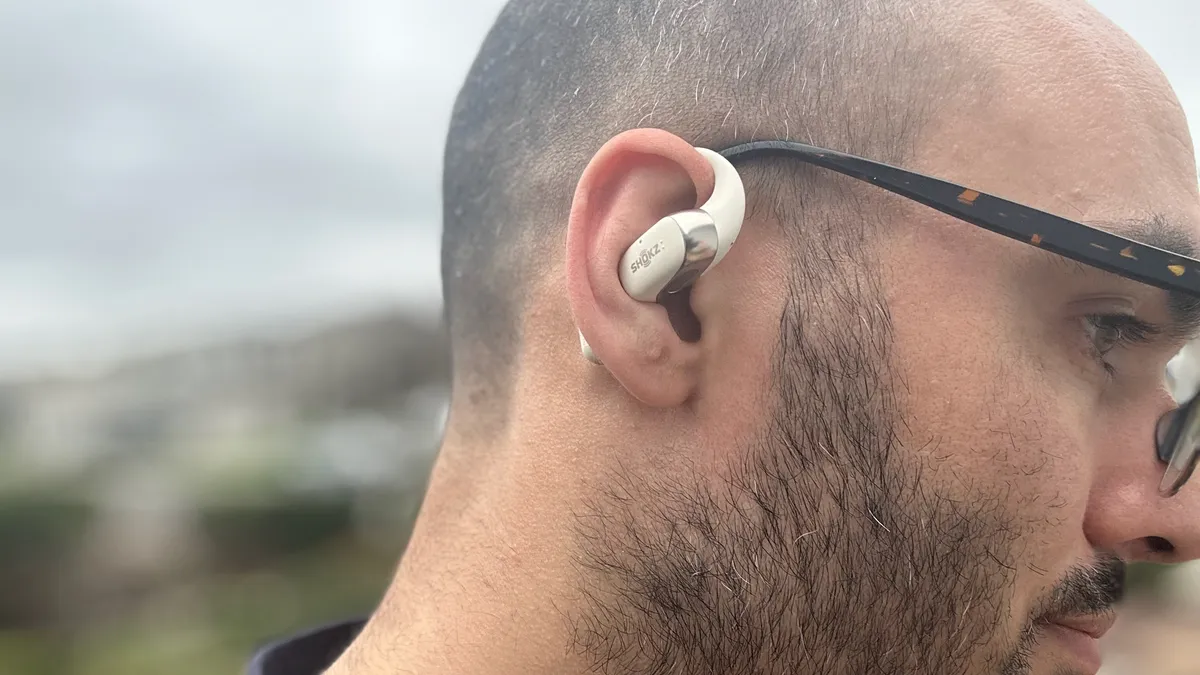 测评者戴着 Shokz OpenFit 2 的照片，耳钩挂在眼镜旁边。