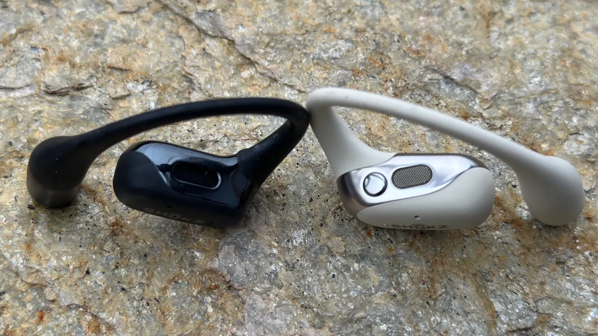 黑色 Shokz OpenFit 和白色 Shokz OpenFit 2 直立在岩石上，对比凸显了第二代型号上的新按钮。