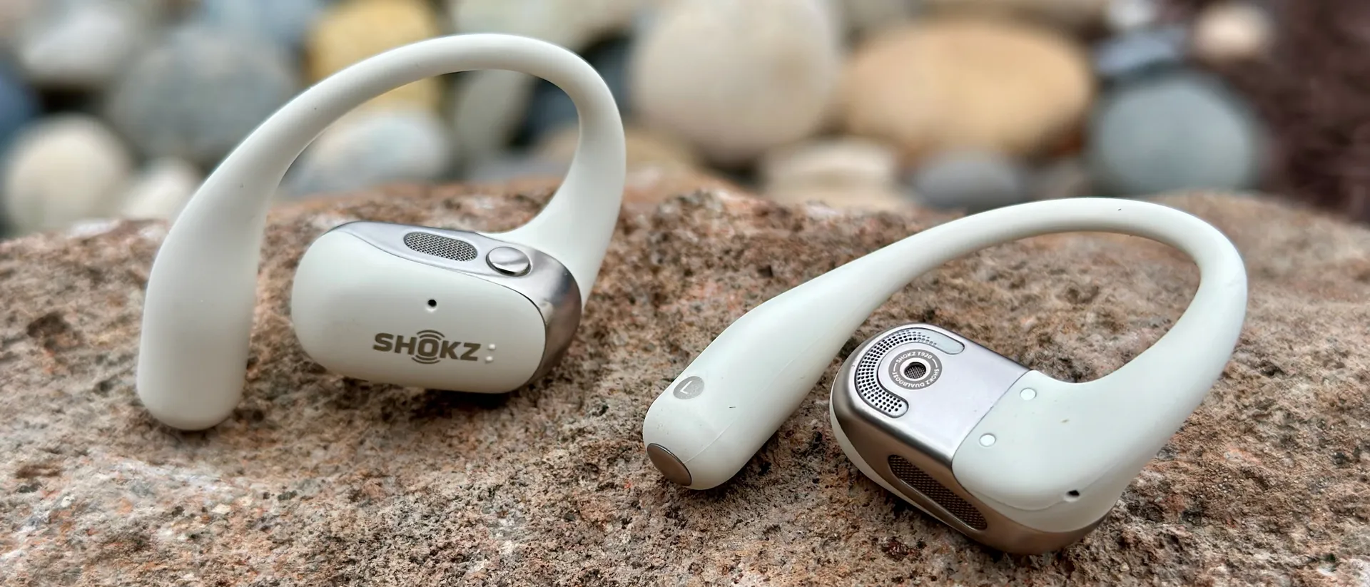 Shokz OpenFit 2 耳塞的特写，一个面向相机，显示徽标和顶部按钮/扬声器格栅，另一个背面朝上，显示主扬声器格栅、麦克风孔和耳钩。