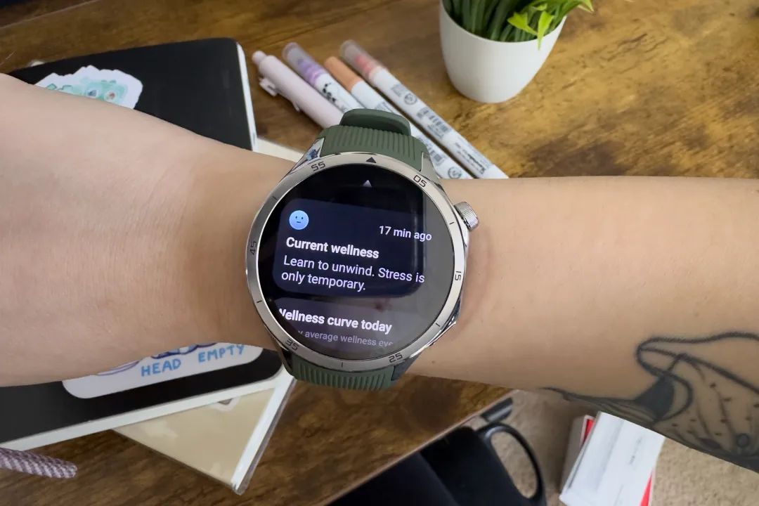 高级评测员 Victoria Song 手腕上佩戴 OnePlus Watch 3 的照片。照片中展示了 360 度身心磁贴，描绘了 Victoria 当前的健康状况，并配上一张面无表情的蓝色表情符号。上面写着“当前健康状况：学会放松。压力只是暂时的。”背景中有一株植物、笔记本和钢笔。