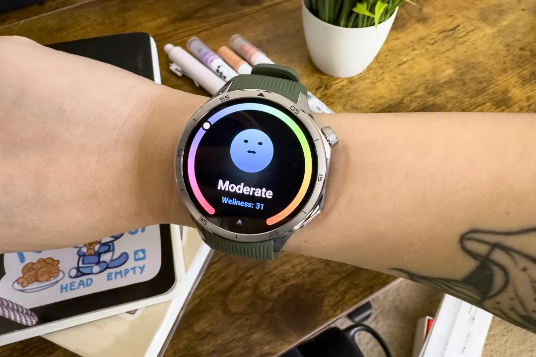 高级评测员 Victoria Song 手腕上的 OnePlus Watch 3 近照。在她较小的手腕上，这款手表看起来略大。屏幕上显示一个面无表情的蓝色表情符号，表示她的健康状况一般。在背景中，你可以看到桌子上的笔记本、笔和桌上植物。