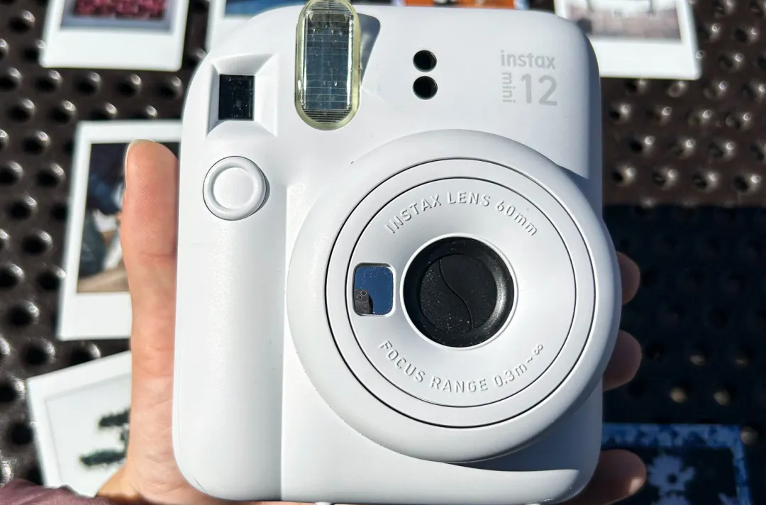 富士 Instax Mini 12 相机的特写，其镜头结构周围环绕着背景中打印的照片。