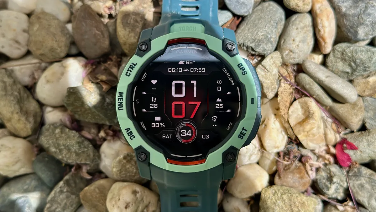 Garmin Instinct 3 50mm AMOLED 坐落在鹅卵石和小岩石之上，红白相间的表盘显示各种数据，如时间和日期、天气、海拔、剩余电池寿命和最大摄氧量。