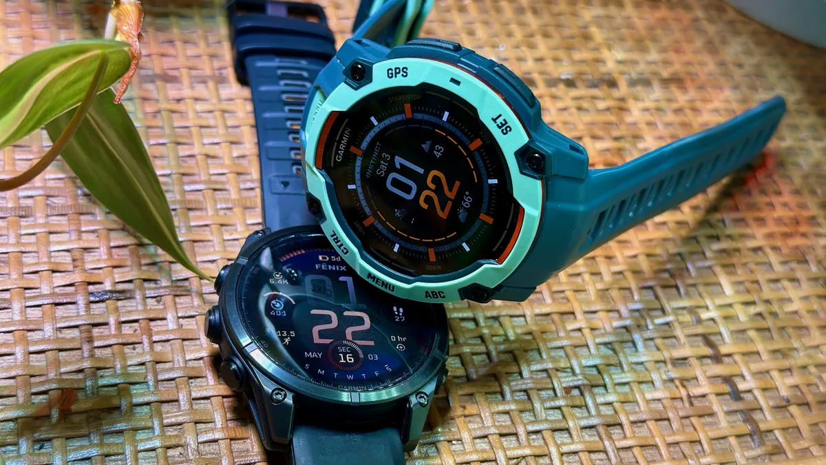 Garmin Instinct 3 50mm AMOLED 侧放在 Garmin Fenix 8 AMOLED 上方，两款手表都位于茅草木架子上方。