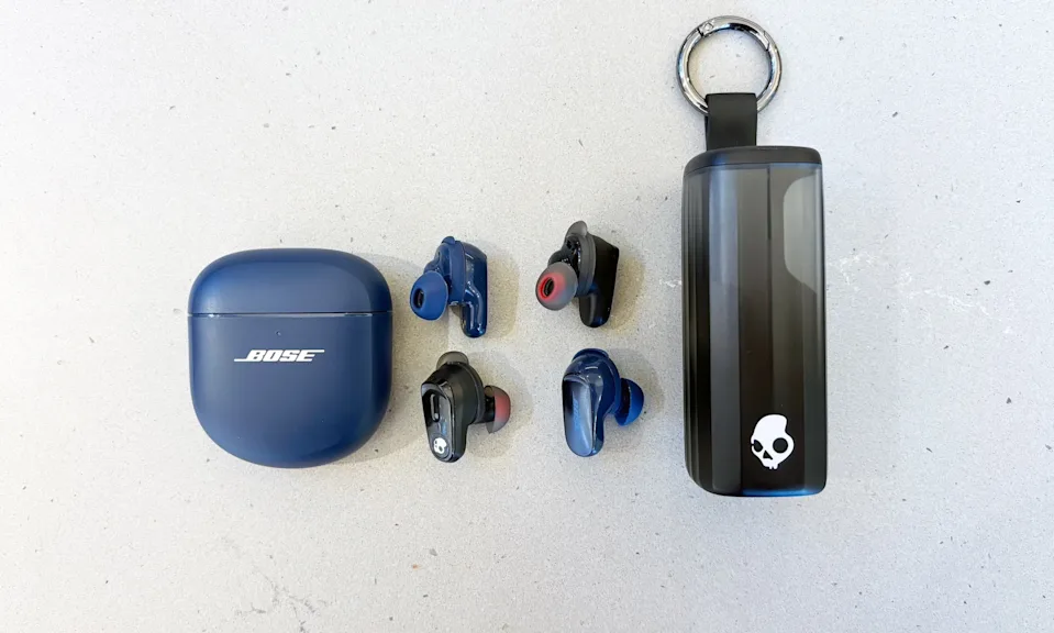 两副无线耳机——Bose QuietComfort Ultra 耳塞和 Skullcandy Method 360 ANC 耳塞——并排摆放在深灰色的台面上。耳机位于两副耳机的充电盒之间。Bose 的耳机为海军蓝，Skullcandy 的耳机为黑色。