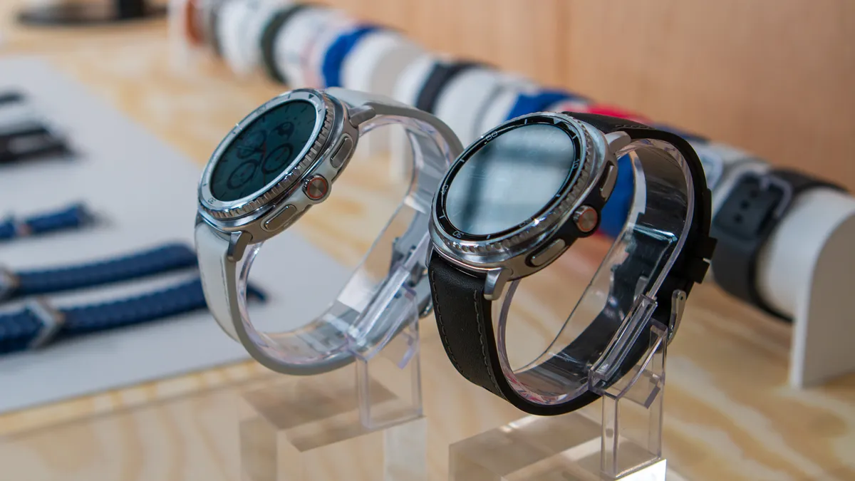 在三星媒体预览活动上，白色和黑色的三星 Galaxy Watch 8 Classic 被放置在桌子上的透明支架上。