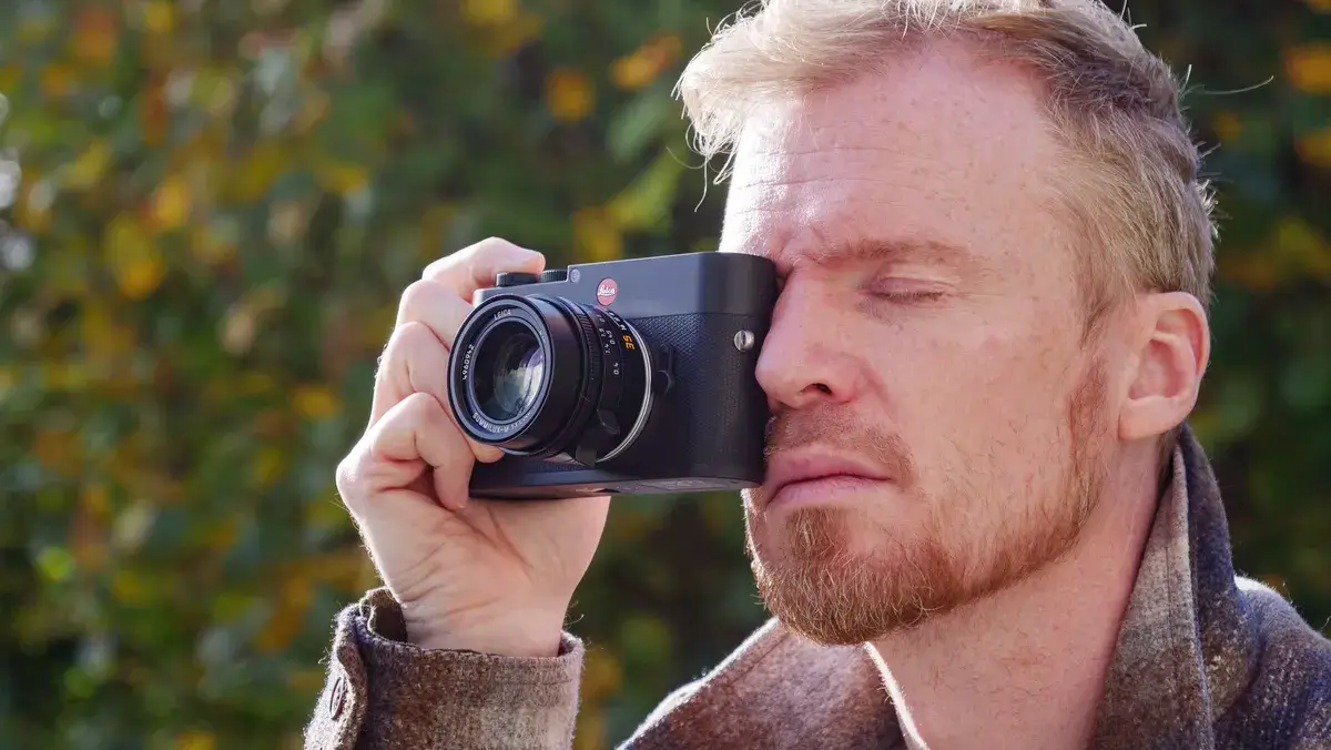 一名男子将徕卡 M EV1 相机的取景器举到眼前，背景是秋叶